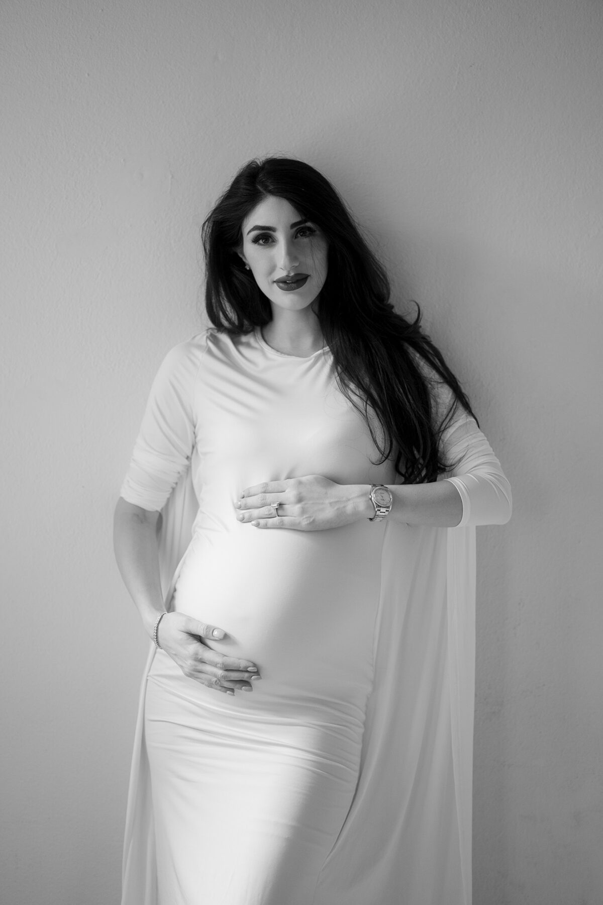 Babybauch Fotoshooting in Wien – Schwarzweißporträt einer schwangeren Frau mit langem, hellem Kleid und Cape, stehend vor einer Wand, mit den Händen auf dem Bauch – elegante Fine Art Fotografie von Shira Rachel Photography