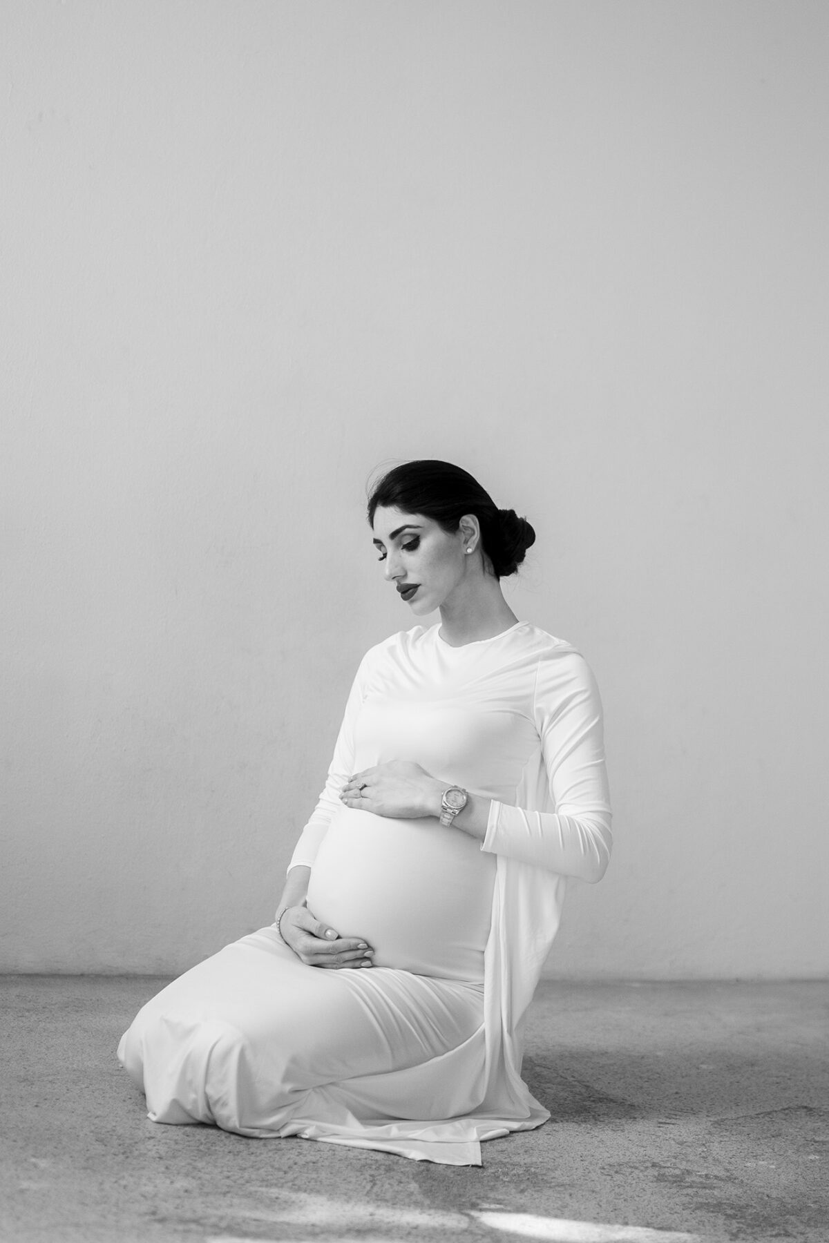 Babybauchfotos in Wien – Schwarzweißaufnahme einer schwangeren Frau in einem fließenden weißen Kleid, kniend mit ruhigem Blick nach unten und Händen auf dem Bauch – minimalistisches Fine Art Porträt von Shira Rachel Photography