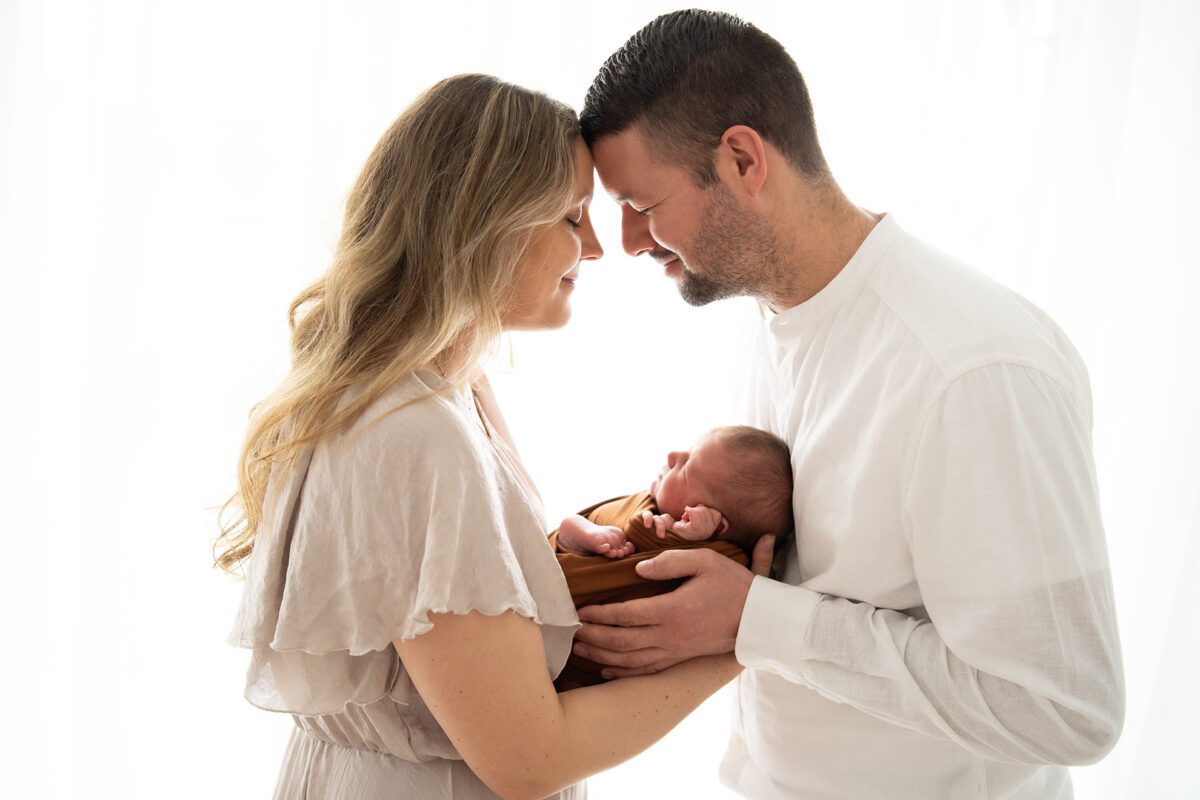 Eltern halten ihr Neugeborenes liebevoll – emotionales Familien-Fotoshooting in Wien. Sie stehen vor einer weißen Kulisse und das Licht strahlt hinein.