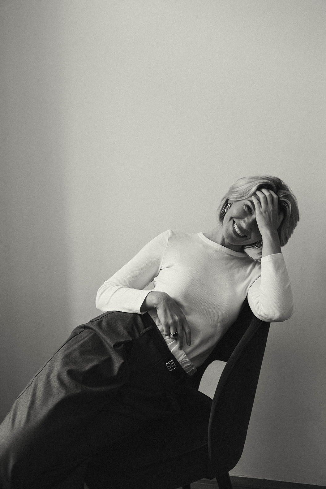 Blonde kurrzhaarige Frau, Portrait der Fotografin in Wien, Shira Rachel, in weißem Oberteil und dunkler Hose sitzt entspannt in einem modernen Stuhl vor neutralem Hintergrund. Ein spontanes Lächeln und die monochrome Bildsprache verleihen dem Portrait Tiefe und Charakter.