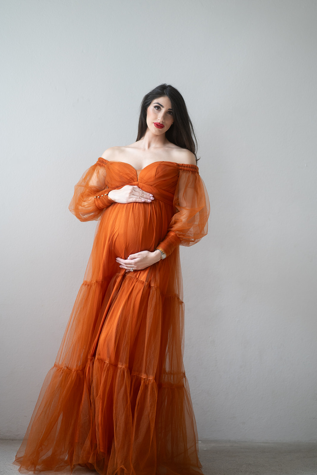 Babybauchfotos Wien. Schwangere Frau in einem eleganten, transparenten orangefarbenen Kleid bei einem professionellen Babybauchshooting in Wien. Sie steht vor einer weißen Wand und hält liebevoll ihren Babybauch – ein stilvolles Beispiel für hochwertige Schwangerschaftsfotografie.