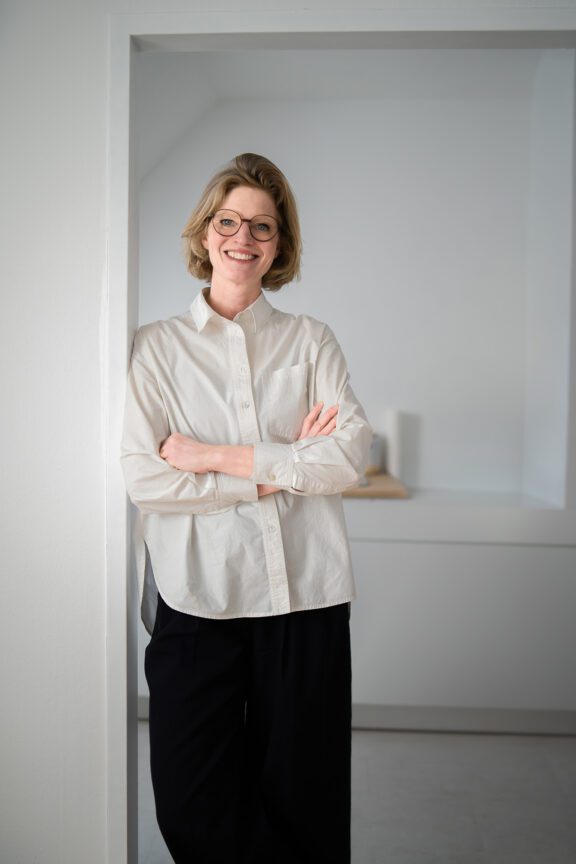 Businessporträt einer Frau mit weißem Hemd und verschränkten Armen in einem minimalistischen Wiener Innenraum – fotografiert von Shira Rachel Photography.