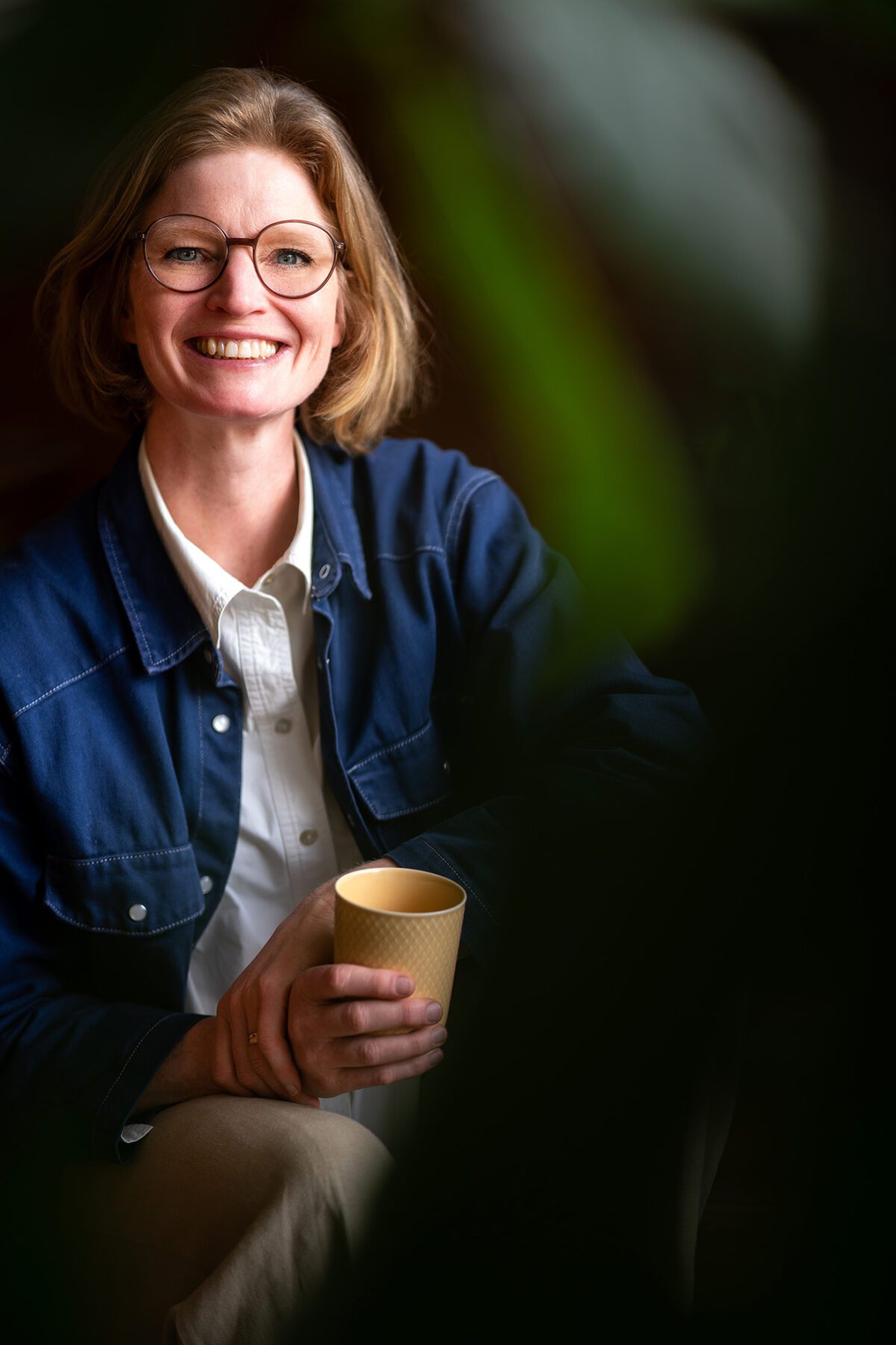 Businessporträt einer lächelnden Person mit Brille, weißem Hemd und blauer Jacke, mit gelber Tasse in der Hand – fotografiert von Shira Rachel Photography in einem natürlichen Setting in Wien.