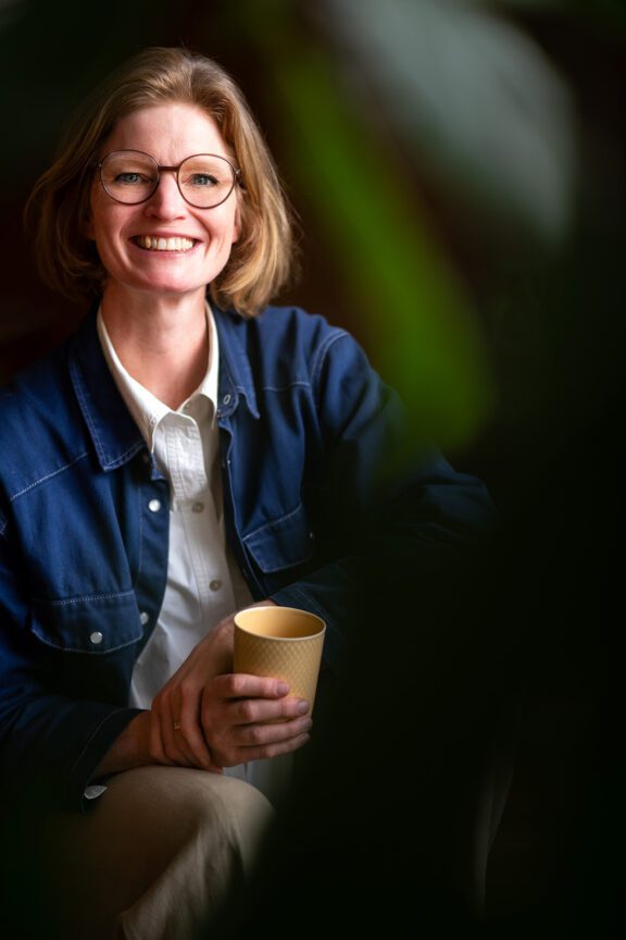 Businessporträt einer lächelnden Person mit Brille, weißem Hemd und blauer Jacke, mit gelber Tasse in der Hand – fotografiert von Shira Rachel Photography in einem natürlichen Setting in Wien.