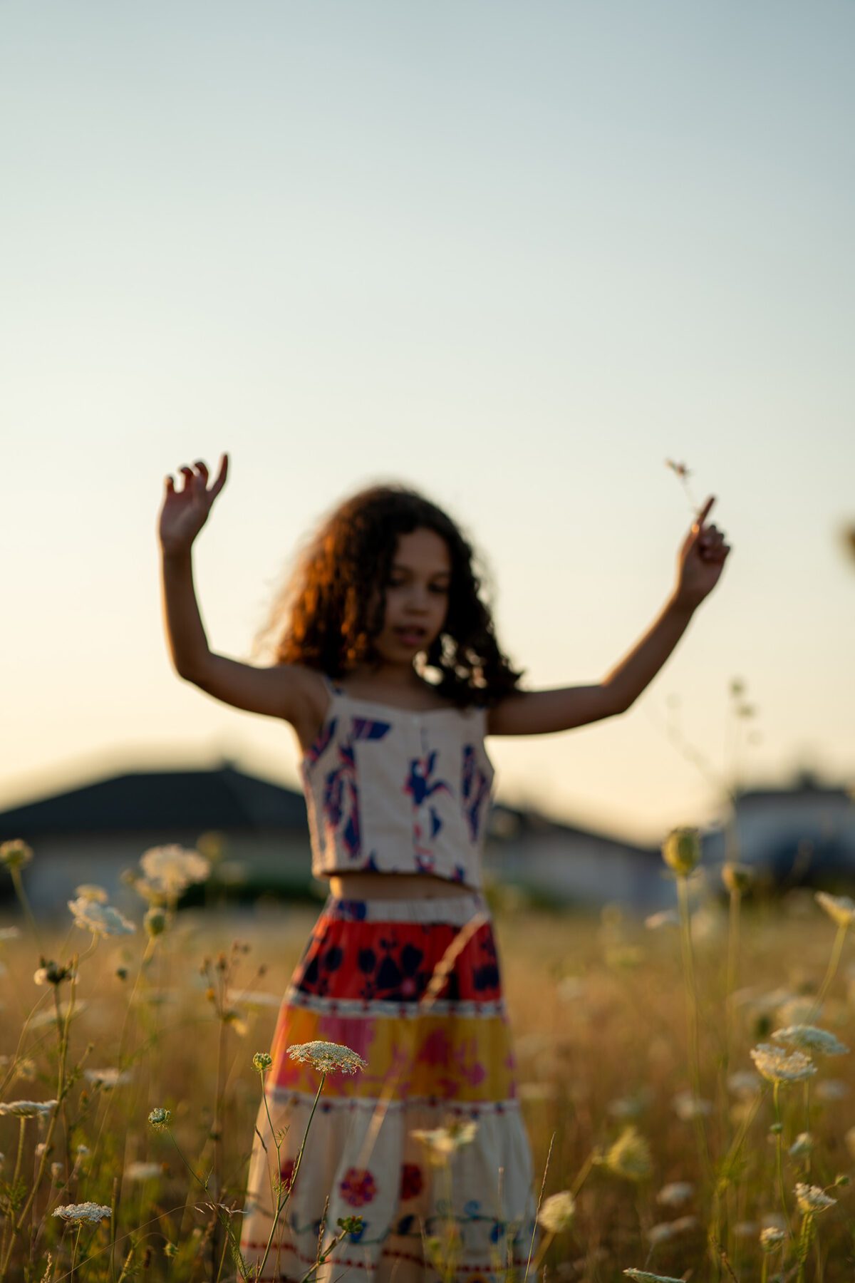 Kind mit lockigem Haar und buntem Outfit steht im Wildblumenfeld bei Sonnenuntergang und hält Blumen in beiden Händen – fotografiert von Shira Rachel Photography in der Nähe von Wien.