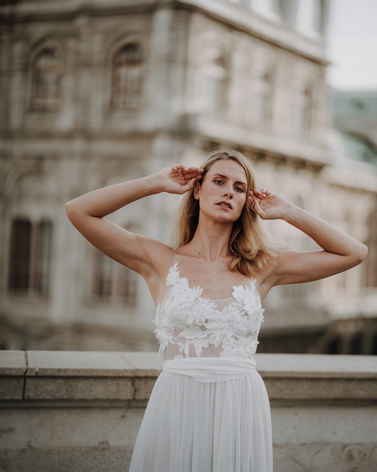 Eine Braut in einem weißen Kleid mit floralen Applikationen posiert auf der Albertinaplatz Terrasse in Wien. Vor der Kulisse klassischer Architektur und weichem Licht entsteht ein ausdrucksstarkes Porträt, das Romantik, Bewegung und urbane Eleganz vereint – aufgenommen von einem Hochzeitsfotografen aus Wien.