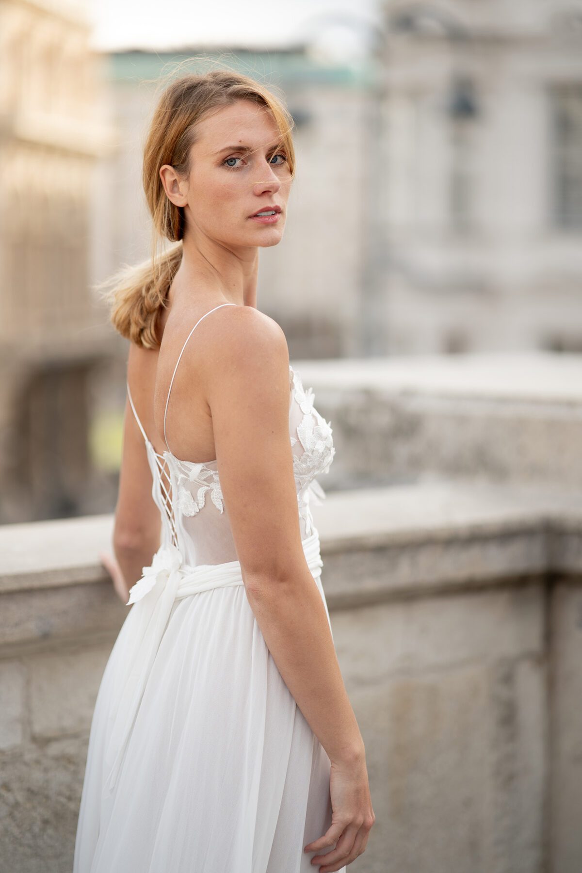 Eine Braut in einem weißen Spitzenkleid mit zarten Trägern steht vor einer historischen Steinbalustrade in Wien. Sie blickt über die Schulter in die Kamera, während das sanfte Licht der goldenen Stunde ihre Silhouette umrahmt. Die Szene strahlt zeitlose Schönheit und romantische Eleganz aus – festgehalten von einem Hochzeitsfotografen aus Wien.