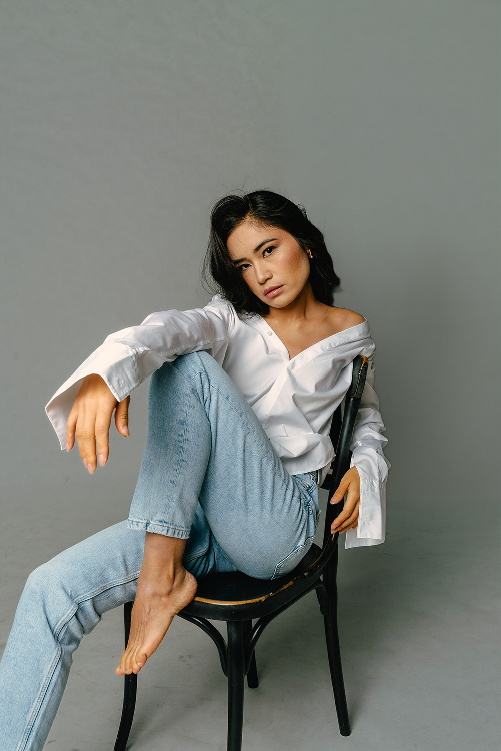 Natürliches Studio-Porträt einer Frau in weißem Hemd und Jeans, entspannt auf einem Stuhl sitzend. Moderne, minimalistische Ästhetik, fotografiert von Shira Rachel Photography.