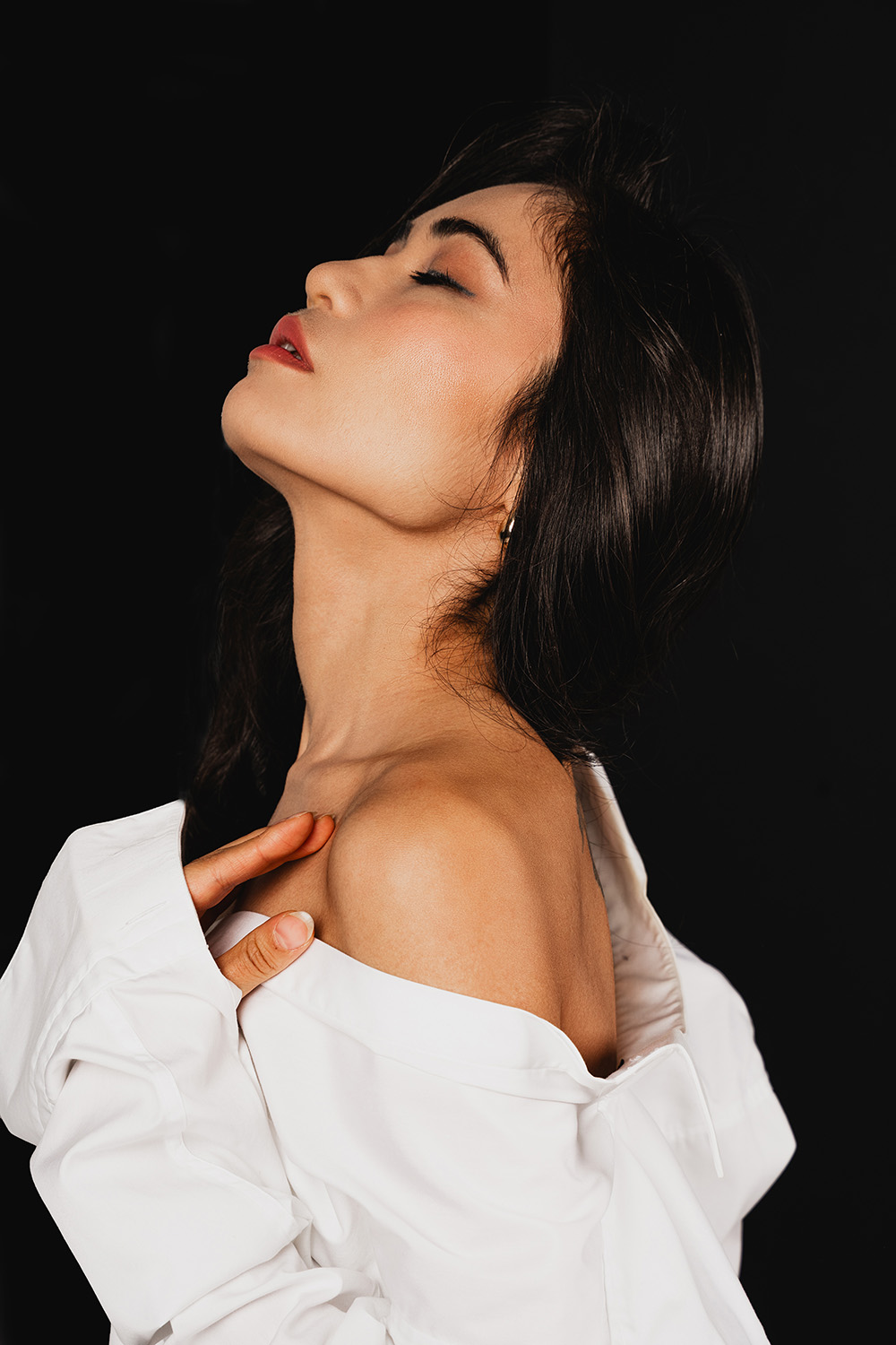 Elegantes Profilportrait einer Frau mit geschlossenen Augen und offener weißer Bluse vor schwarzem Hintergrund, fotografiert von Shira Rachel Photography