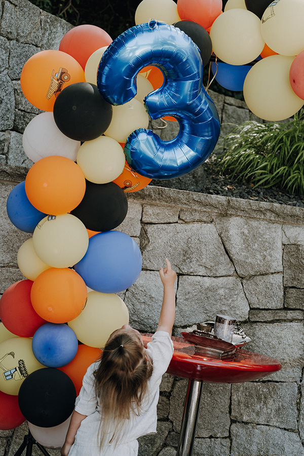 Kind zeigt begeistert auf eine große blaue „3“ vor einer bunten Ballonwand bei einer fröhlichen Feier, liebevoll dokumentiert von einem Familien Fotograf Wien beim Kindergeburtstag.