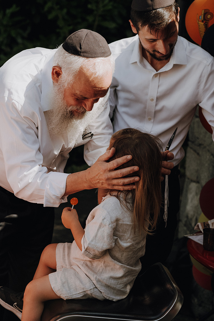 Rabbiner legt einem Kind bei einer upsherin Feier im Freien die Hände auf den Kopf des Geburtstagskind und segnet es, während ein weiterer Erwachsener mit einer Schere bereitsteht – ein besonderer Moment, einfühlsam festgehalten von einem Kinderparty Fotograf Wien.