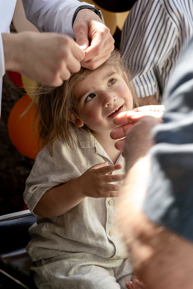 Kind sitzt während einer festlichen Feier auf einem Stuhl und wird liebevoll von mehreren Erwachsenen frisiert, ein besonderer Moment authentisch eingefangen von einem Eventfotograf Wien.