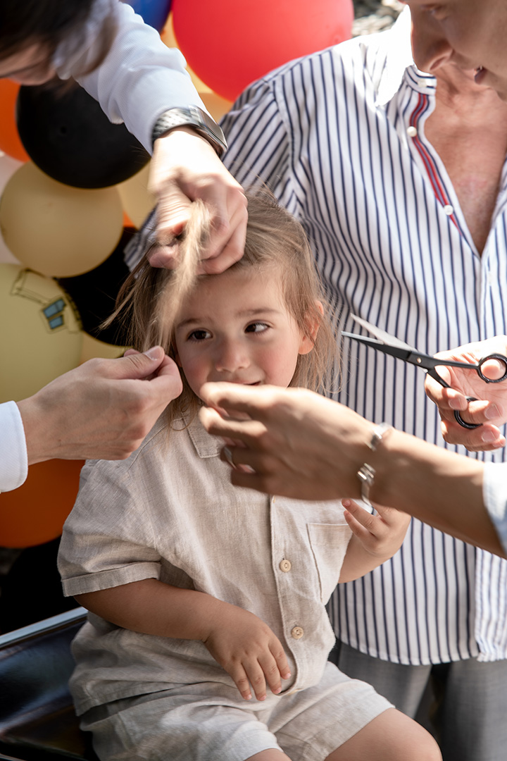 Kind bekommt bei einer Feier im Freien einen Haarschnitt, umgeben von mehreren Erwachsenen und farbenfrohen Ballons, authentisch dokumentiert von einem Eventfotograf Wien.