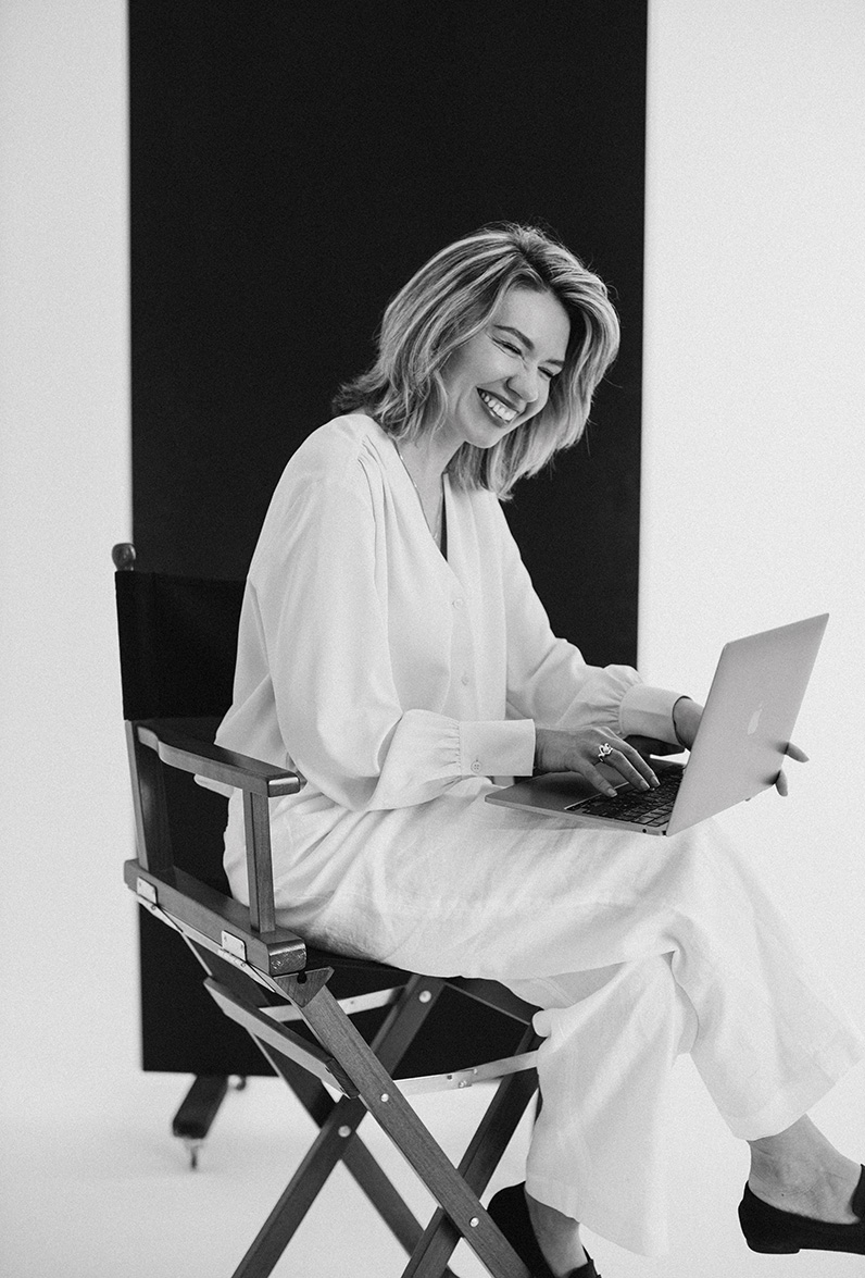Shira Rachel, Fotografin aus Wien, lacht herzlich, während sie im Regiestuhl sitzend an ihrem Laptop arbeitet, Schwarz-Weiß-Porträt im Studio